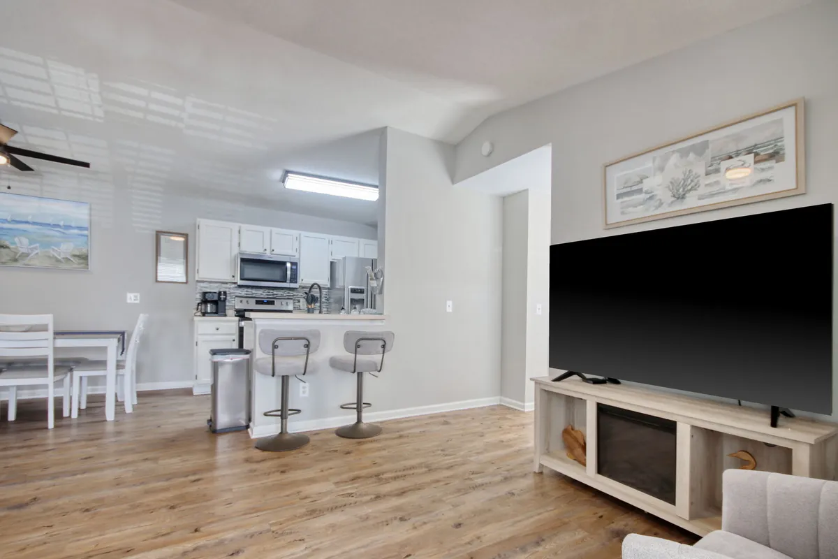 This image captures an open interior layout where a large television is positioned near a kitchen area and a dining table, all set within a single, inviting space. The view showcases how the living, dining, and kitchen seamlessly blend together, providing a vibrant space for socializing, eating, and entertainment. The room is filled with natural light, highlighting both communal and leisure features.