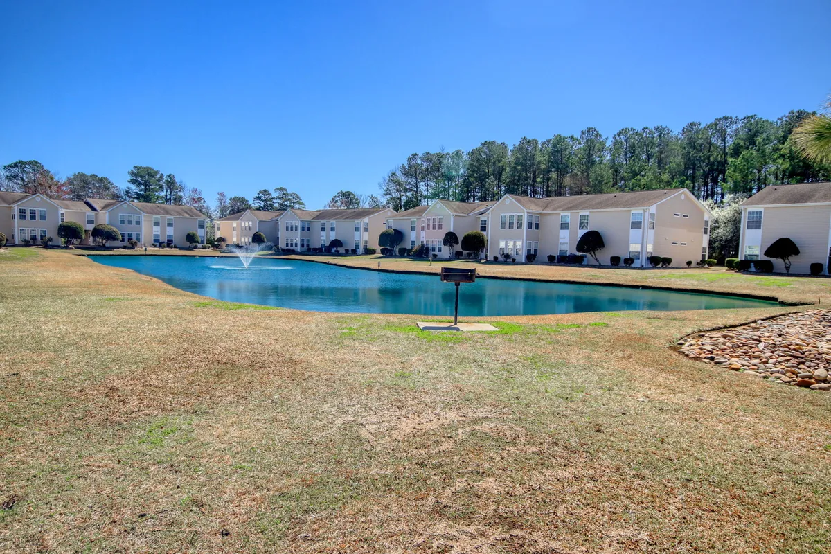 An inviting exterior view featuring a tranquil pond with a fountain set against landscaped grounds and residential buildings. The area looks ideal for picnicking, relaxing by the water, or enjoying a peaceful community event outdoors.
