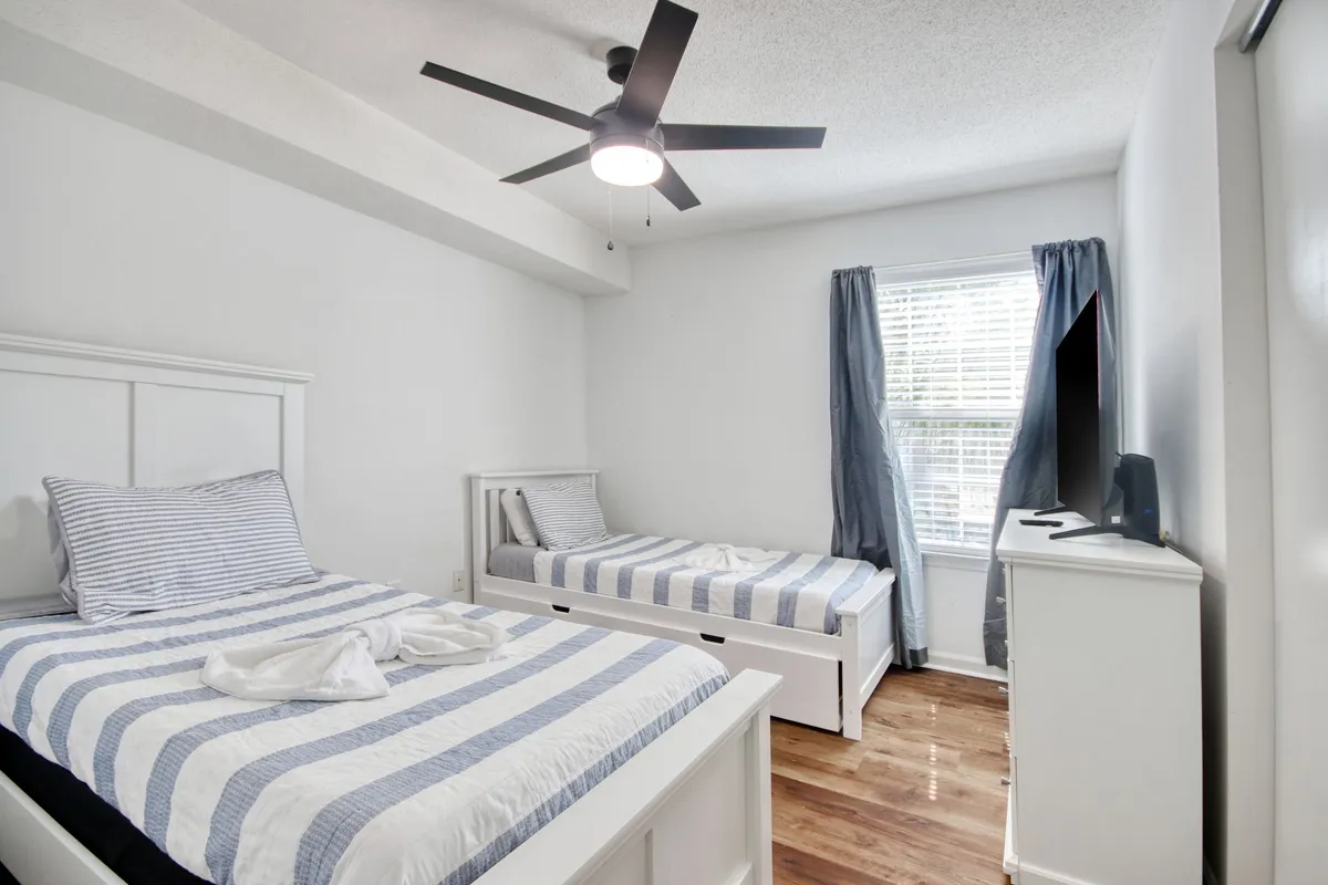 A bright twin bedroom with two beds covered in striped bedding, a window letting in natural light, a ceiling fan for comfort, and a dresser with a television. The setup is perfect for families or friends sharing a room.