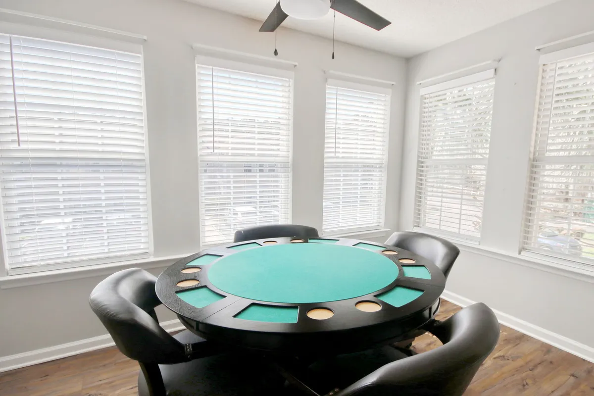 A round poker table is the centerpiece of this image, flanked by comfortable black leather chairs and natural light filtering through window blinds. The setup hints at game nights, friendly competitions, and social gatherings in a welcoming atmosphere.