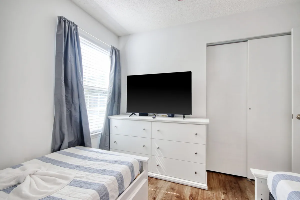 A cozy bedroom featuring a bed with striped bedding, a white dresser, a flat-screen TV, and a window with light curtains. The room exudes a serene and peaceful atmosphere, ideal for relaxation after a day of activities.